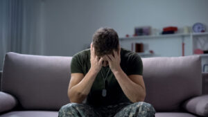 A man in military attire holds his head in his hands while sitting on a couch. Learn how trauma therapy in Arcadia, AZ can offer support in coping with PTSD by contacting a therapist in Arcadia, AZ, and other services today. 