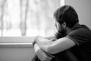 A man sits huddled next to a window. Learn more about how trauma therapy in Arcadia, AZ can offer support in overcoming isolation by searching “trauma counseling Phoenix AZ” or contacting a therapist in Arcadia, AZ today.