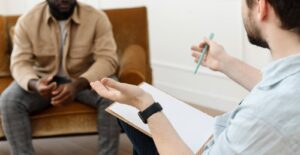 A person with a clipboard and pen gesturing to a man sitting across from him. This could represent the support that trauma therapy in Arcadia, AZ can offer. Learn more about how a therapist in Arcadia, AZ can offer support by searching “trauma counseling phoenix az” today.