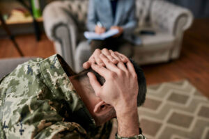 A man in a military uniform holds his head in pain while a person with a clipboard takes notes. This could represent the support a therapist in Arcadia, AZ can offer. Learn more about trauma therapy in Arcadia, AZ and other services by searching “PTSD treatment Scottsdale” today. 